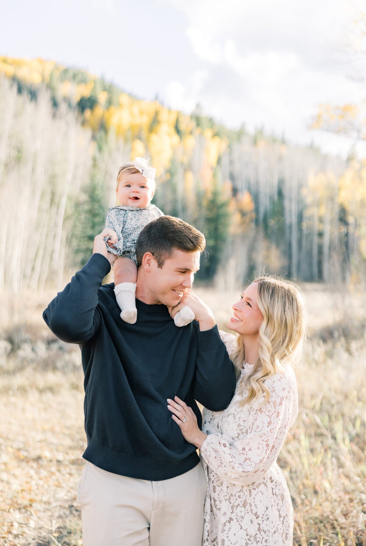The same family of three has their baby on Dads shoulder. The mom is in a white dress and dad is in a dark blue sweater. They are surrounded by mountain fall colors. 