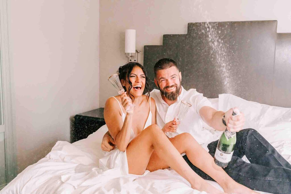 Bride and groom pop a champagne bottle on a bed, to celebrate their engagement.