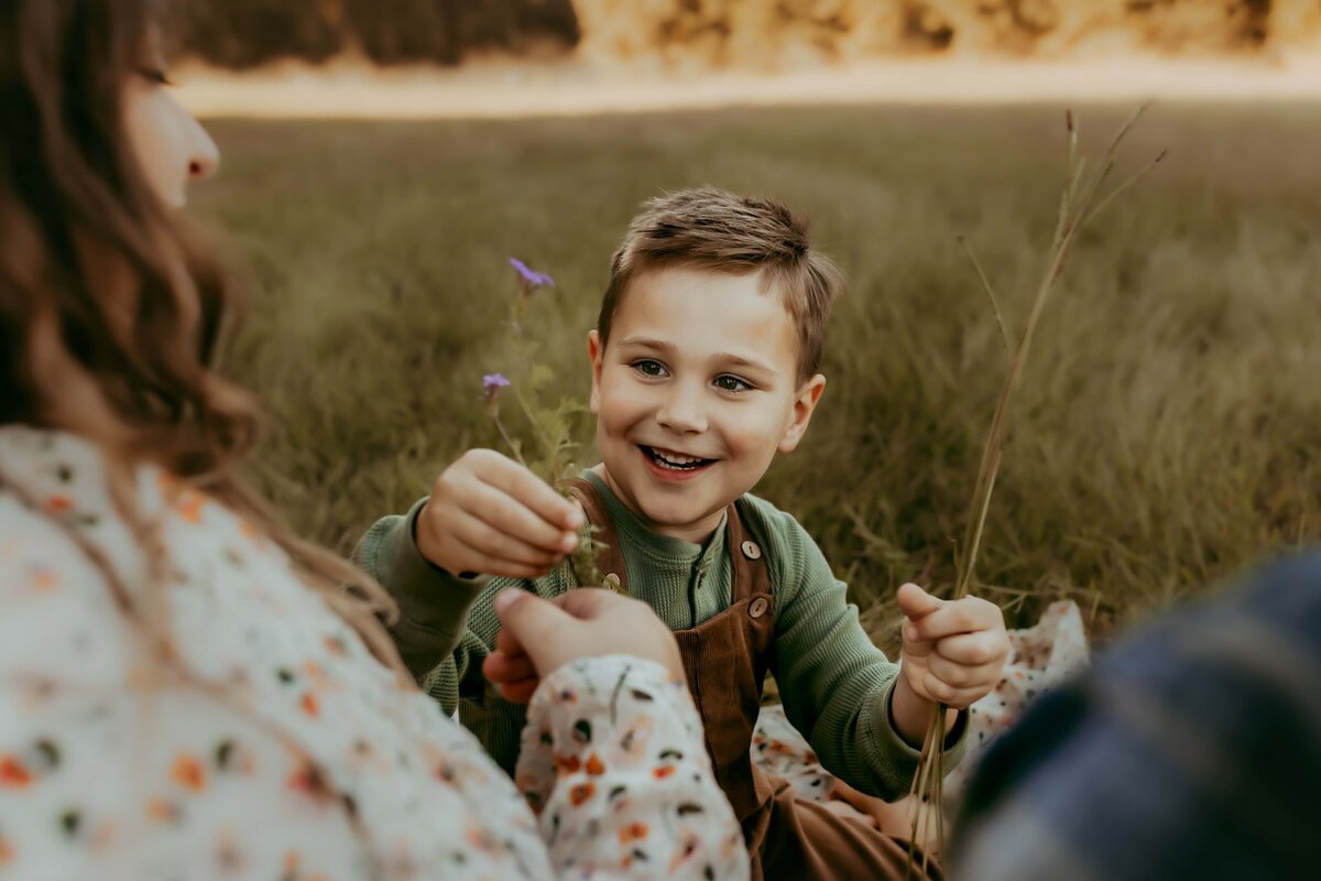 A child in a green shirt and brown overalls sits outdoors on a blanket with an adult, offering a small flower. Capturing this tender moment is a specialty for any family photographer in Warner Robins, GA.