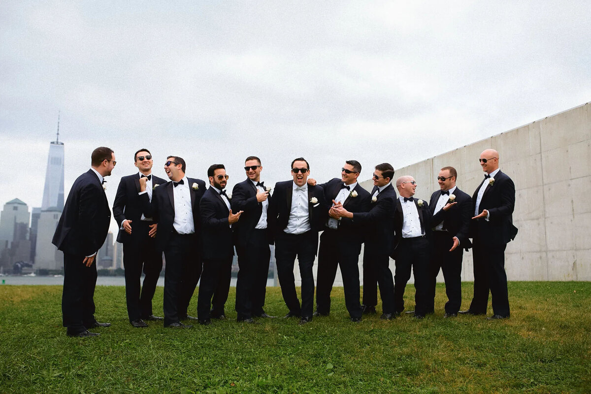 A group of eleven men in tuxedos and sunglasses stand together on grass, laughing and talking, with a city skyline and cloudy sky in the background—captured by an experienced NJ wedding photographer.
