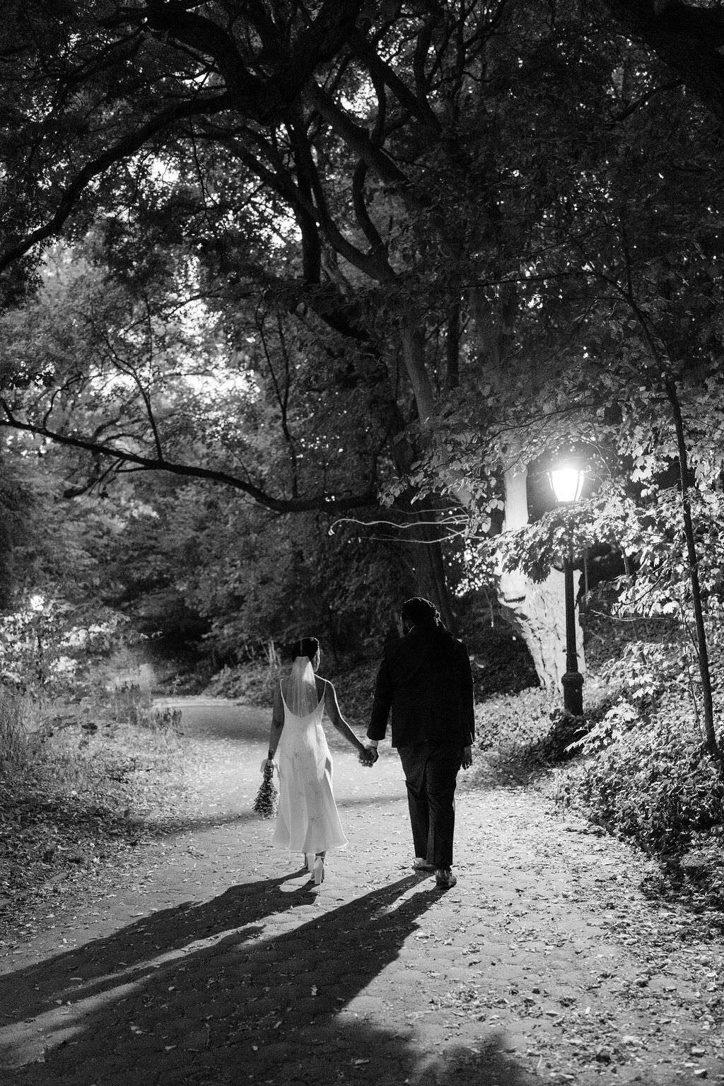 black and white photo of a bride and a groom holding hands and walking on a path in a park