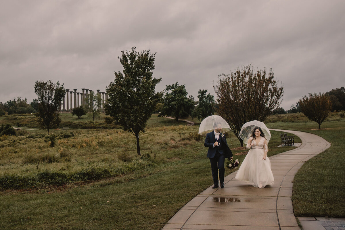 A newlywed couple walking on a sidewalk while holding clear umbrellas over them 
