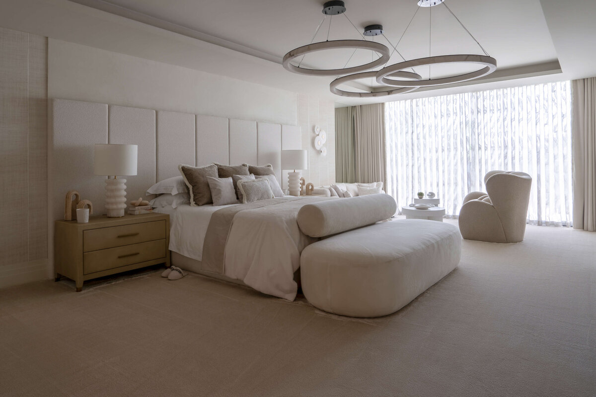 A spacious primary bathroom with a  bright, airy feel. The large, white bed is decorated with neutral accent pillows, with light wood side tables at each side. There is a large white footboard bench, and above the bed is a large, contemporary light fixture. composed of three circles.