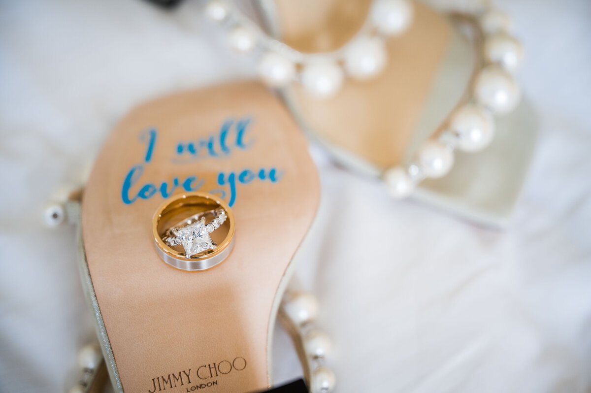 The bottom of a pair of bride's wedding shoes with the words "I will love you" written on them and two wedding rings sitting on them.