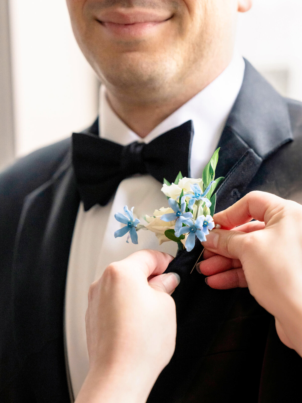 A person in a black tuxedo with a bow tie is having a boutonniere with blue and white flowers pinned to their lapel by another persons hands. The background is softly blurred.