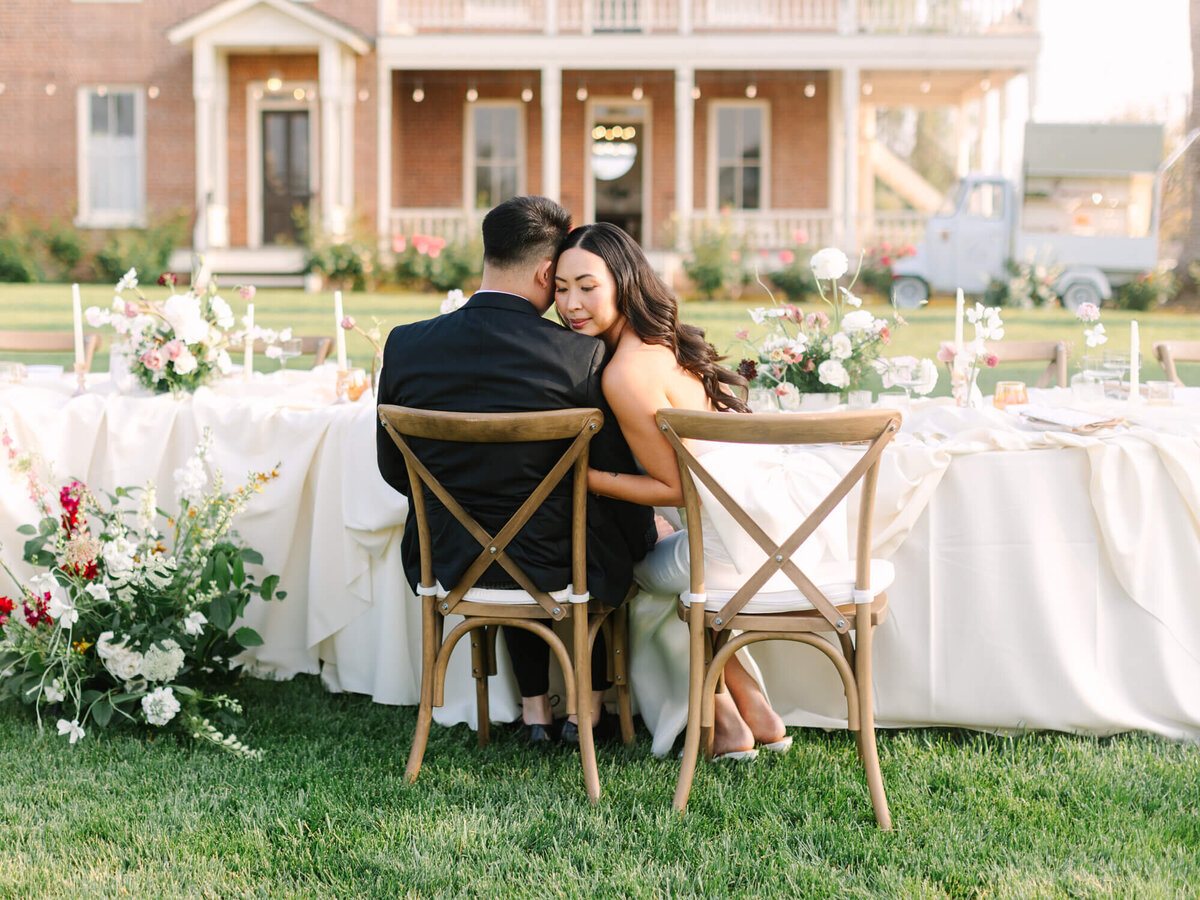 A couple sits closely by an elegantly set outdoor table, surrounded by lush flowers.