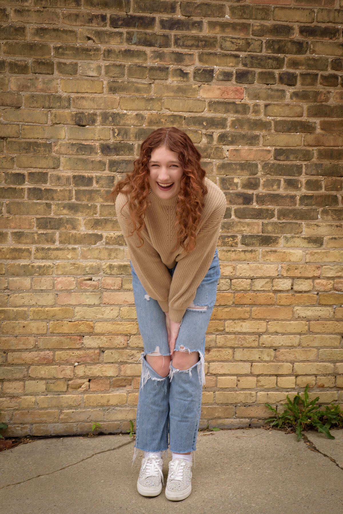 Green Bay East High School senior girl wearing a tan sweater and jeans standing and laughing in front of a tan brick wall in an urban setting in downtown Green Bay, Wisconsin.