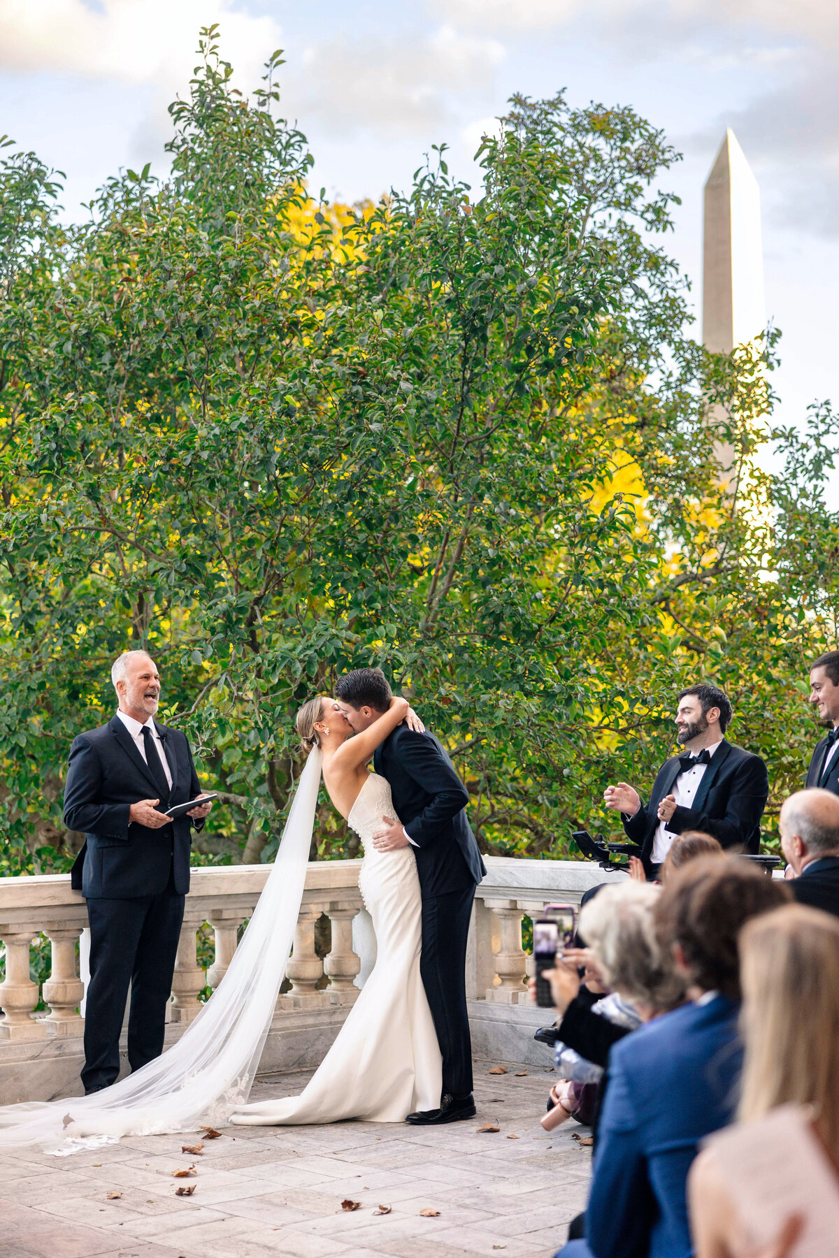 A bride and groom share a kiss during their outdoor wedding ceremony, surrounded by guests and lush greenery. A tall obelisk is visible in the background, and a person officiates the ceremony nearby.