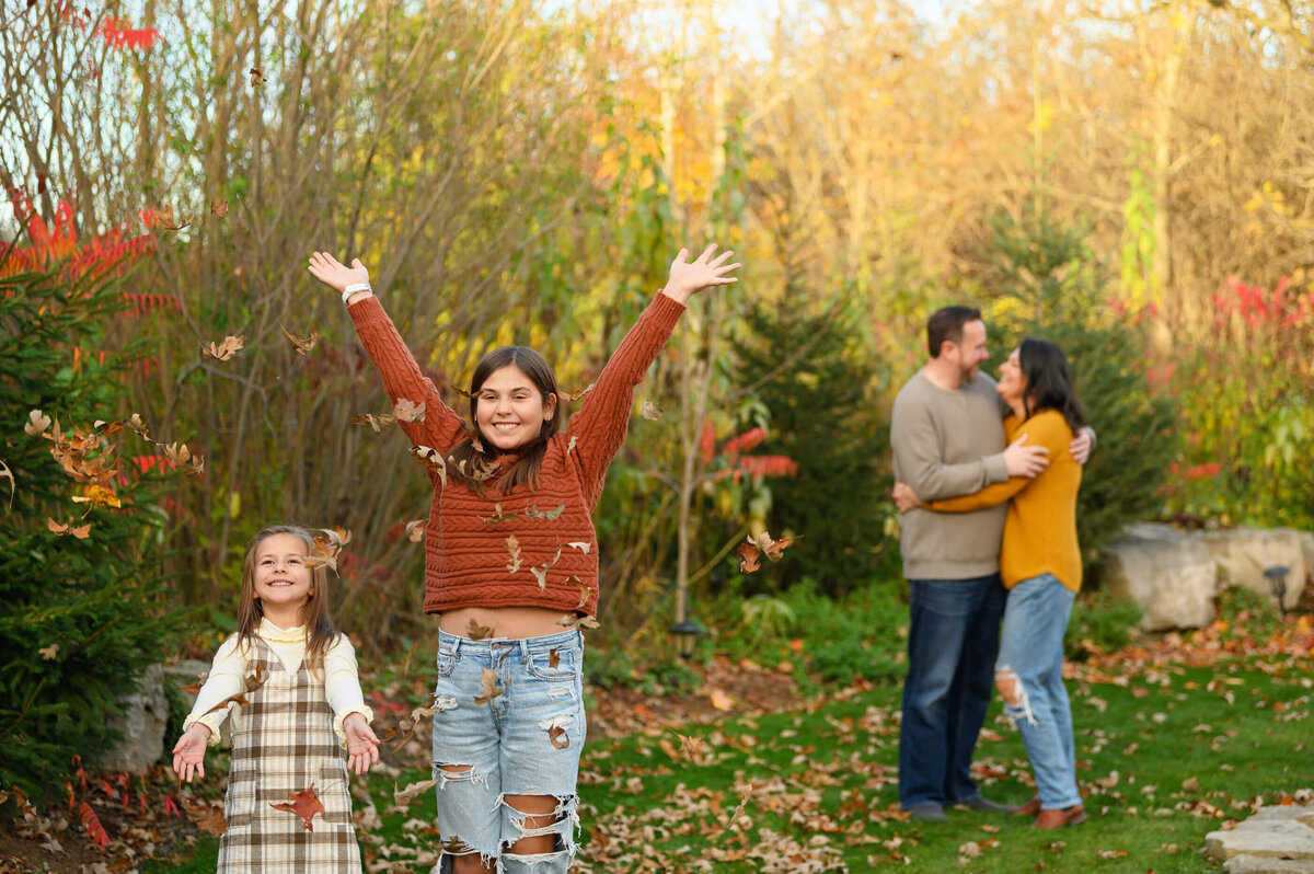 two sisters in the forefront throwing leaves in the hair with mom and dad in the background hugging for their fall family photographer session in Burlington, ON