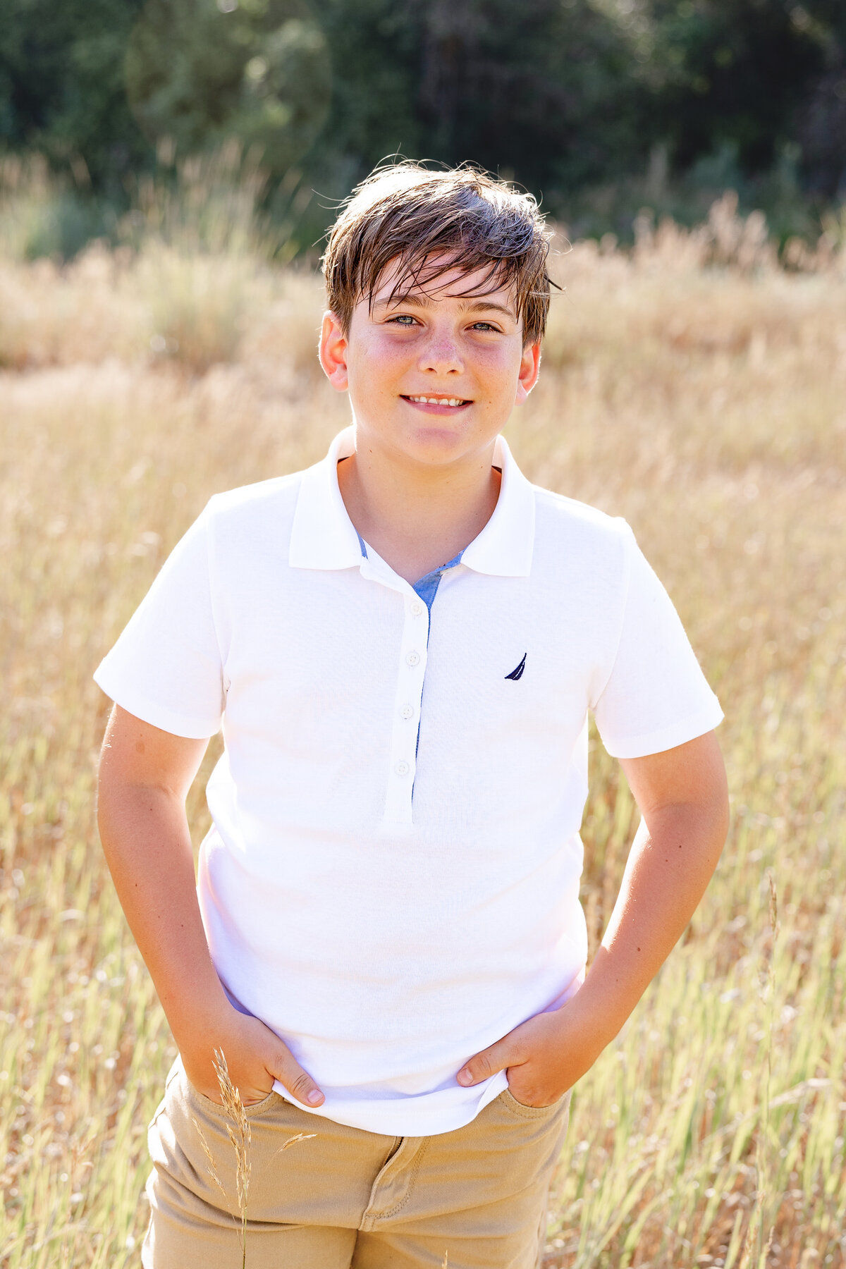 A boy with short brown hair smiles with his hands in his pockets, dressed in a crisp white polo shirt and khaki pants, standing in a golden field.