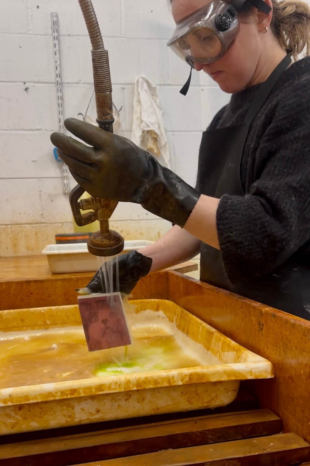An artist wearing gloves and goggles rinses a copper plate in a safety sink during the toner transfer etching process.