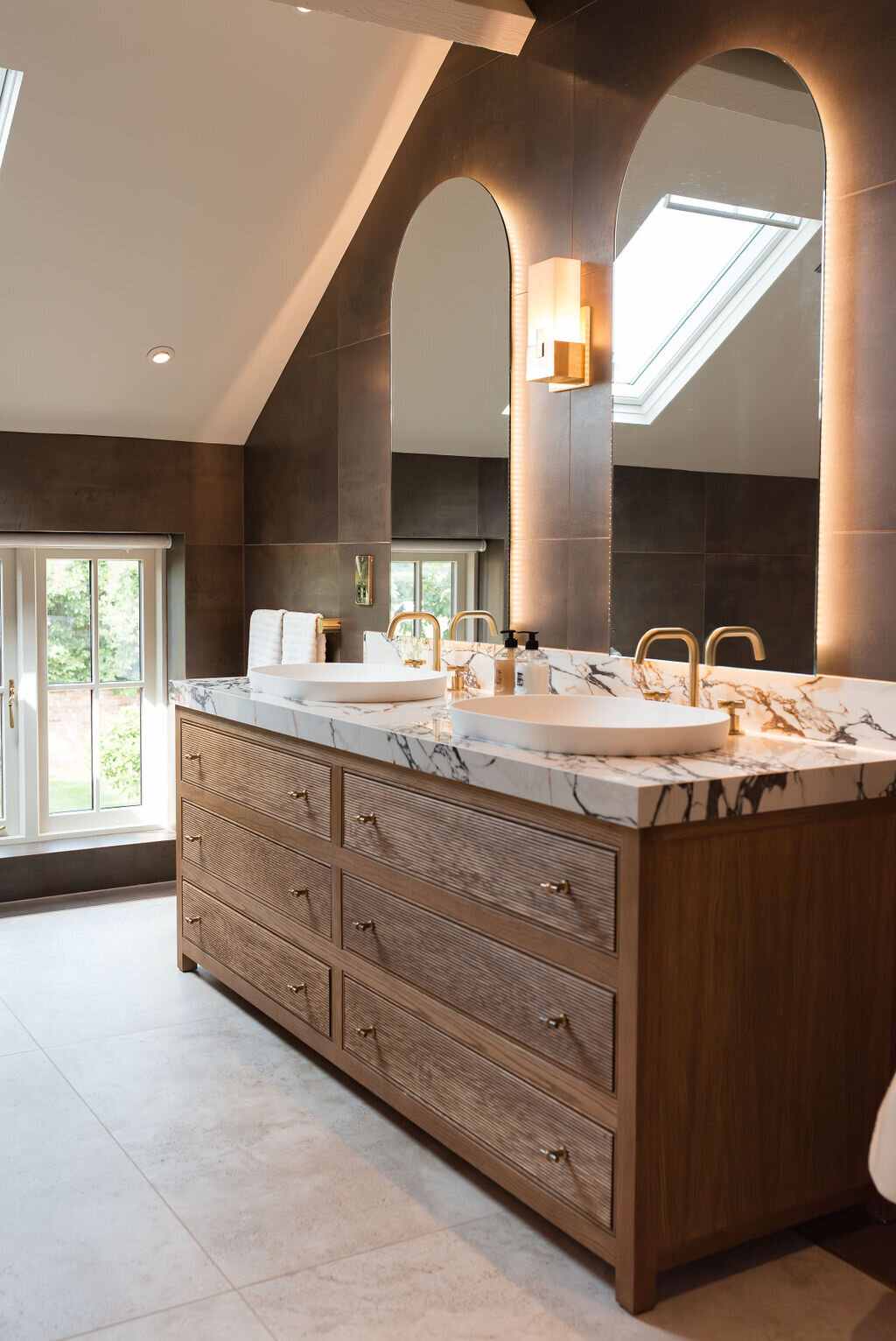 A modern bathroom with a sloped ceiling, featuring a double sink vanity with marble countertops and wooden cabinets. Two arched mirrors are mounted above the sinks, and large windows provide natural light. The room is finished with dark tiled walls and light tiled floors.