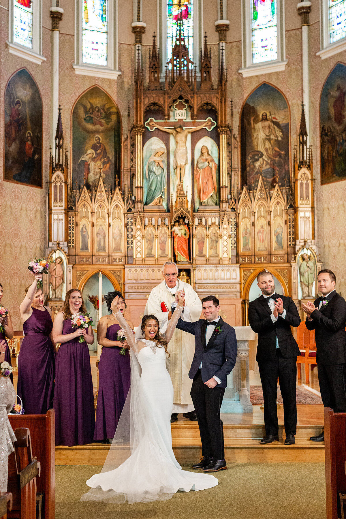 Bride and groom stand hand in hand celebrating in front of an ornate church altar with the bridesmaids in purple dresses and groomsmen in black suits stand on either side, clapping and smiling.