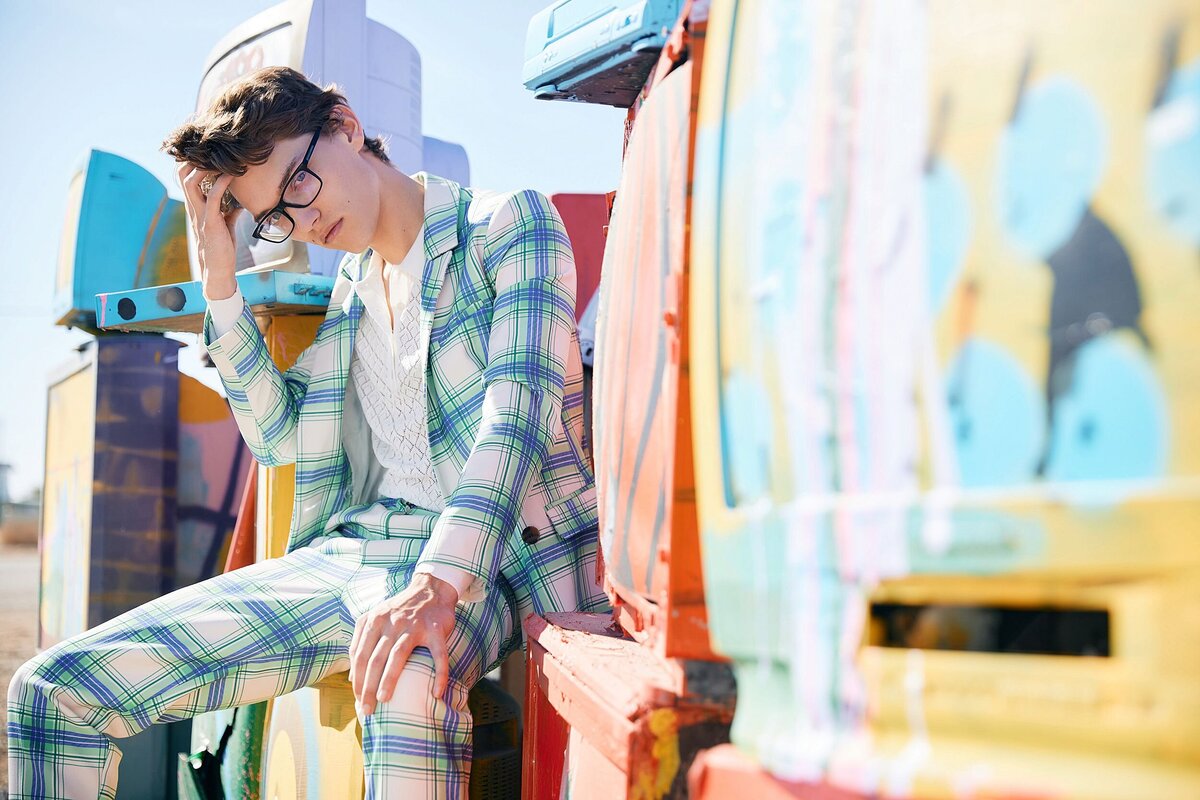 Teen guy wearing a bold and fashionable plaid suit sitting on multiple colorful televisions