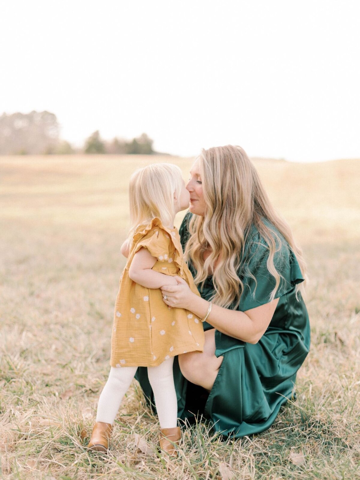 Mother kneels down to kiss daughter in grass field.