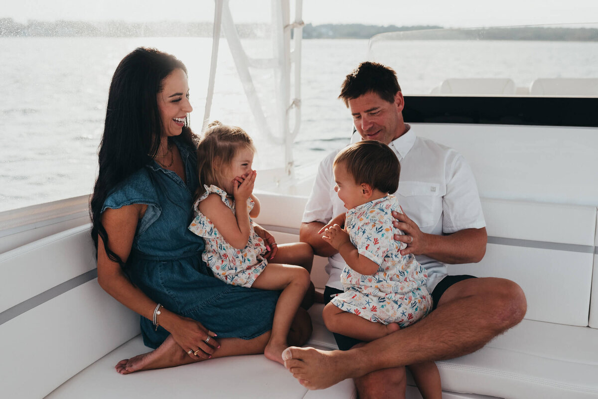 A father and mother sit on their boat during sunset with their two young children facing one another