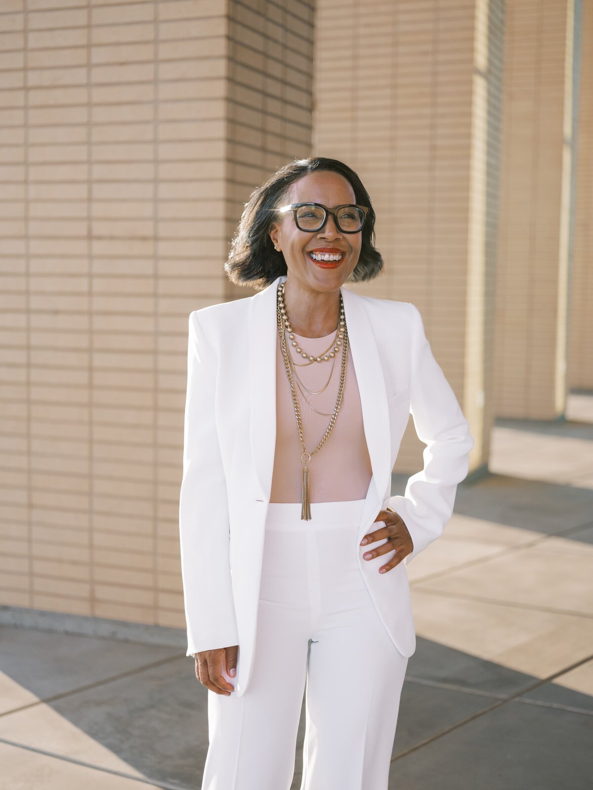 Woman standing in a brick courtyard wearing a white suit ad pink shirt and smiling away from the camera