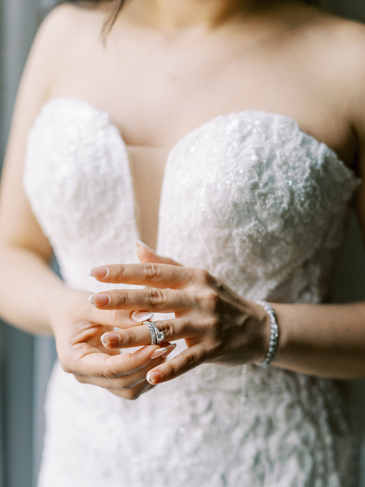 A bride in a strapless white lace wedding dress with a deep neckline, holding her hands together, showcasing a ring and a bracelet at her classic Calgary wedding.