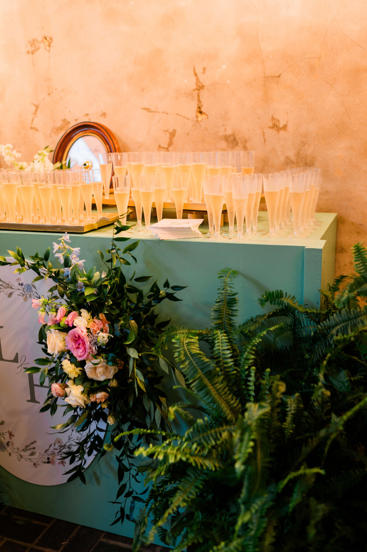 A green table decorated with a floral arrangement and ferns holds numerous filled glasses. Behind the glasses is a round clock against a textured, beige wall.