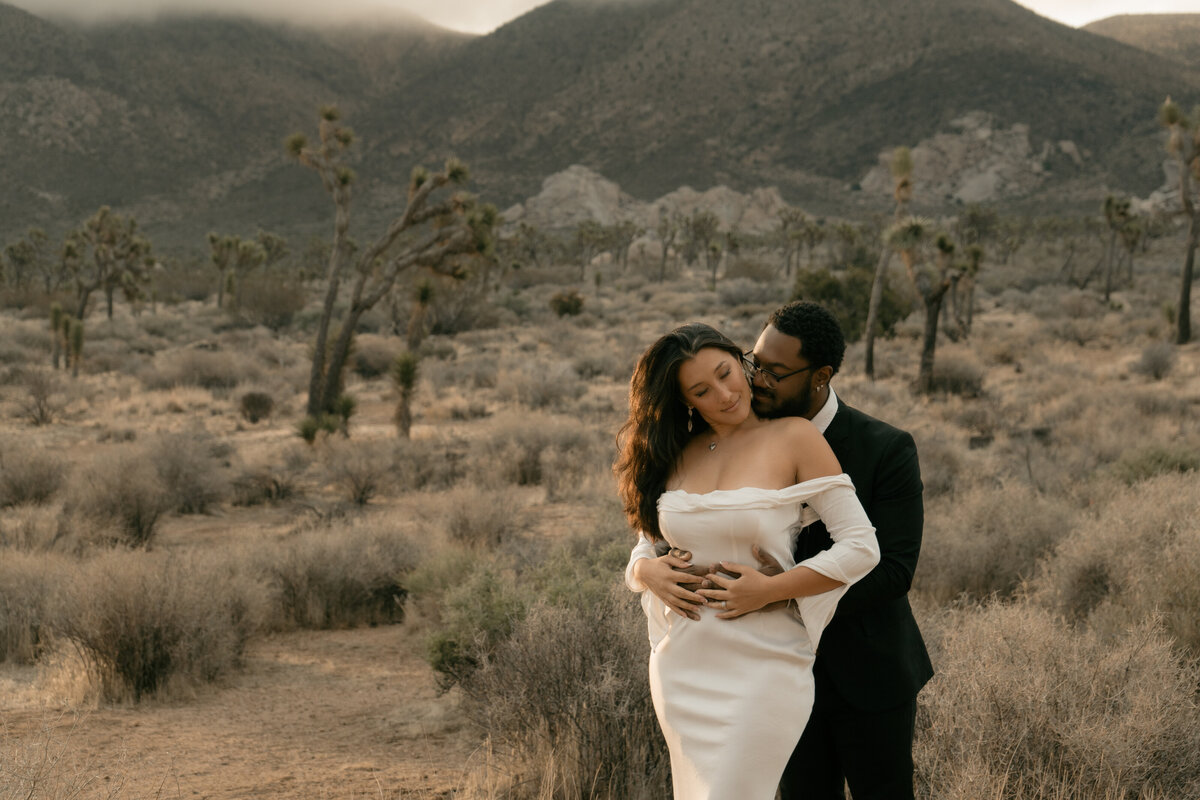Bride and groom at Joshua Tree during sunrise with the groom kissing the bride's neck while embracing her in the desert scenery