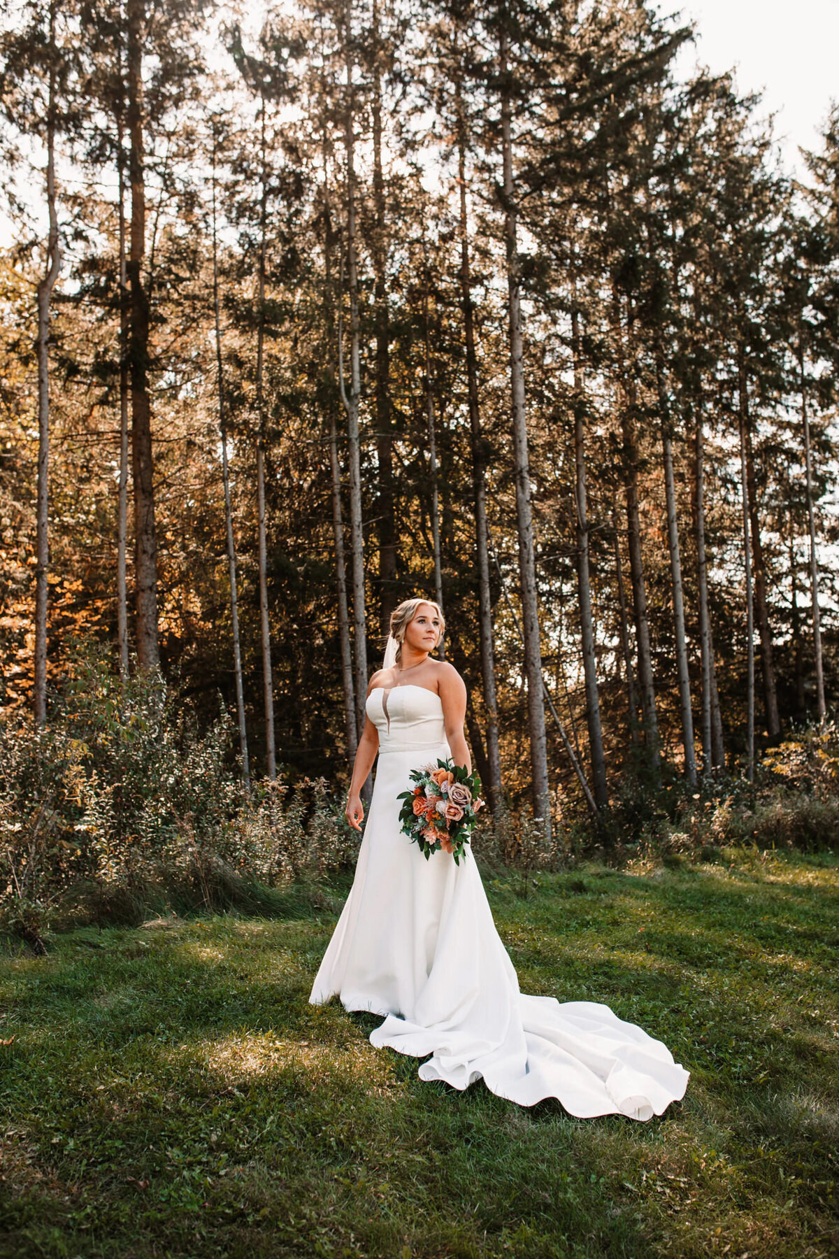 bride stands alone in front of forest while holding flowers down at her side as she gazes into the distance