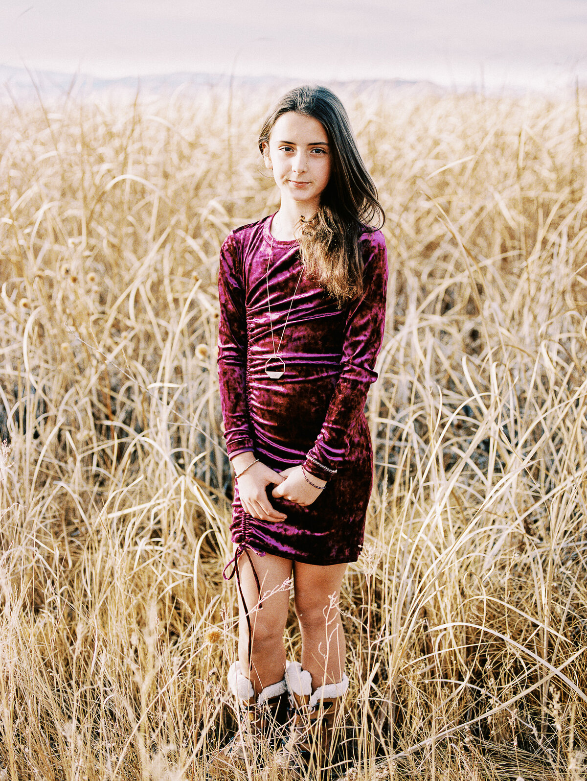 a girl in a maroon dress stands in tall dry grass with mountain in the distance