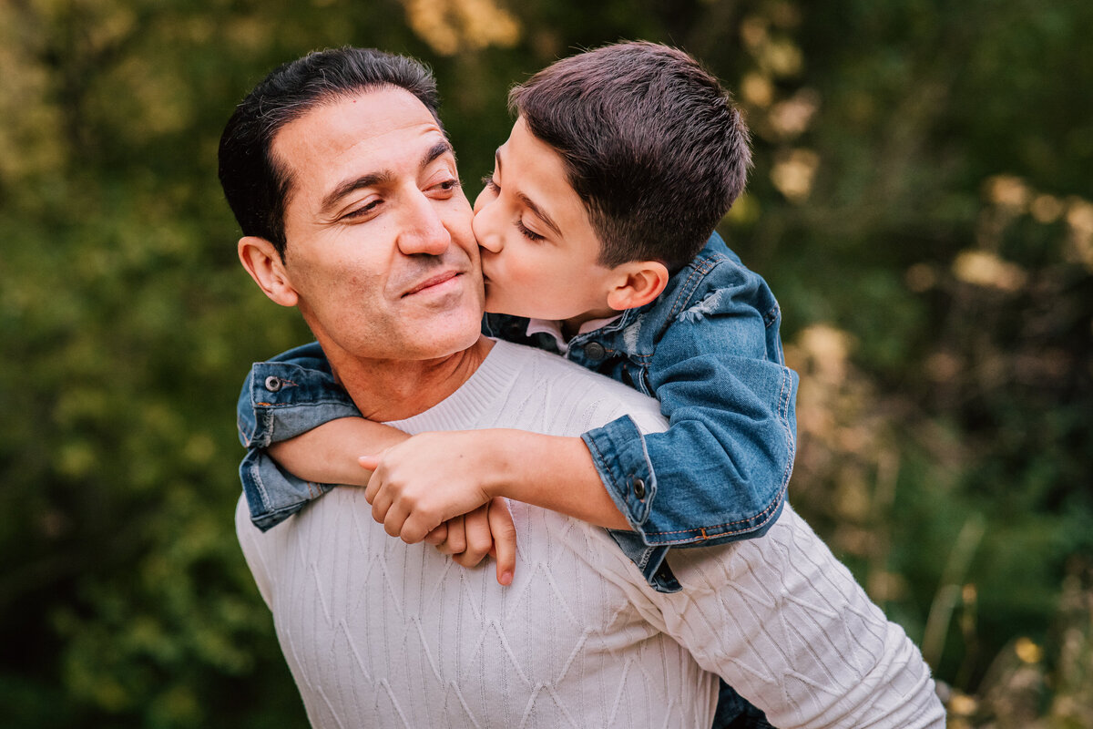 A color photo of a father and son sharing a tender moment outdoors, with the son giving his father a kiss on the cheek while riding piggyback.
