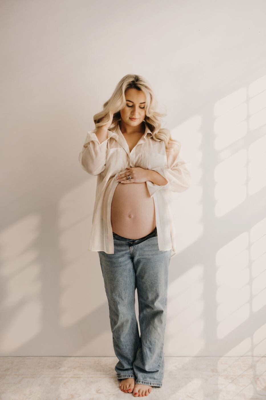 Full-length maternity portrait of a pregnant woman with long blonde wavy hair gently holding her baby bump in a bright studio in Mullingar, Ireland. Professional pregnancy photoshoot