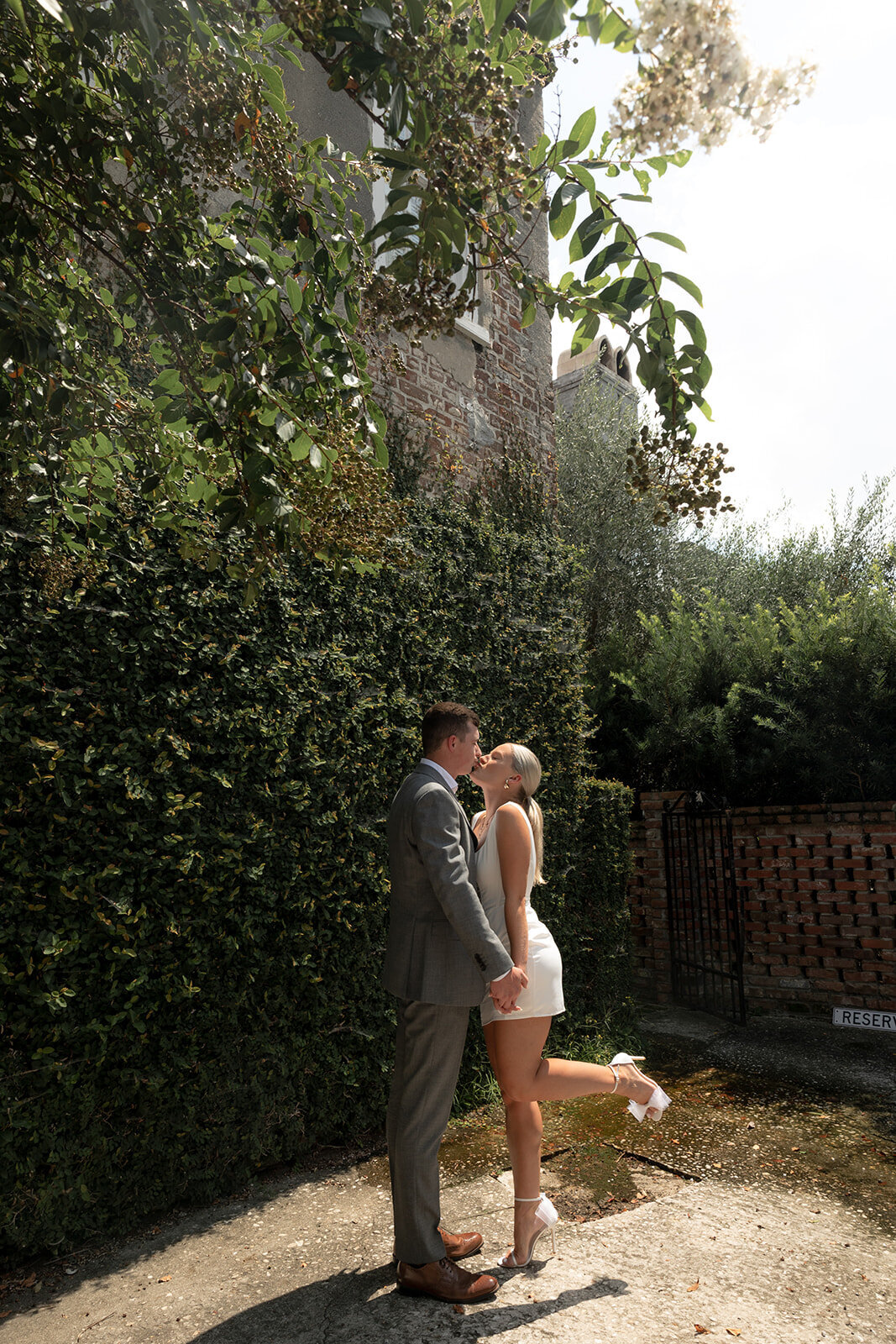 Couple poses for engagement photos. Holding hand while kissing. WOman with one leg up.