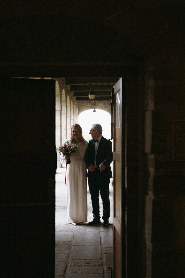 Mariée et son père prêts à entrer dans la cérémonie, capturés à travers une porte. Photo Kamescop, photographe et vidéaste mariage.