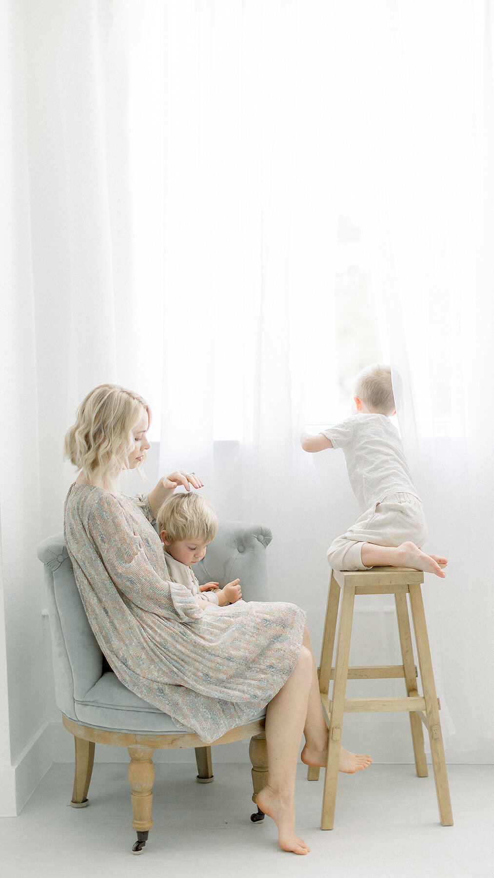 A light filled photo of a mother sitting on a chair in a Dallas photography studio while one of her boys sits on her lap and the other is peeking outside of the window.