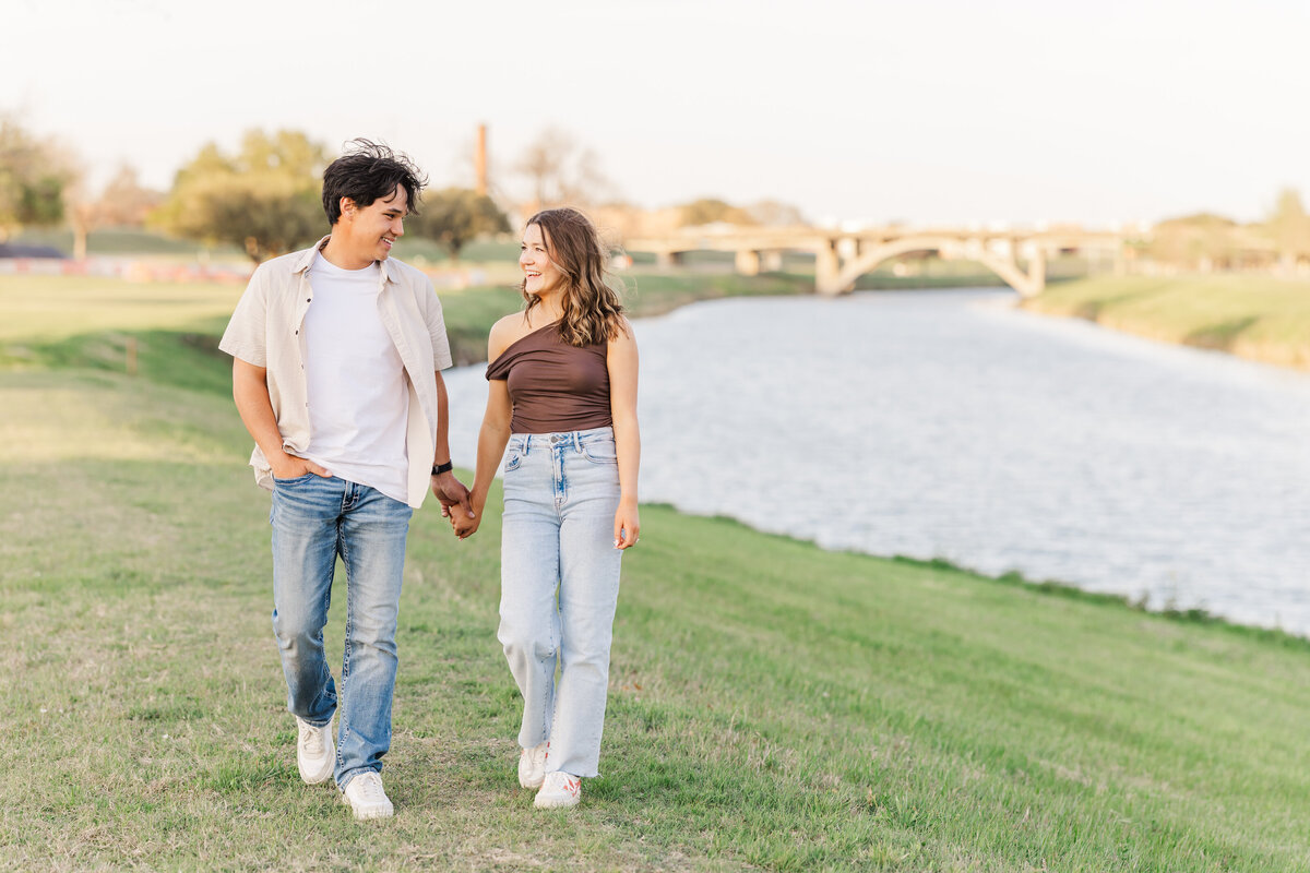 A joyful couple strolls hand in hand along the riverbank during golden hour in Fort Worth. This light and natural engagement portrait captures a relaxed, candid moment with soft sunlight and a scenic Texas backdrop.