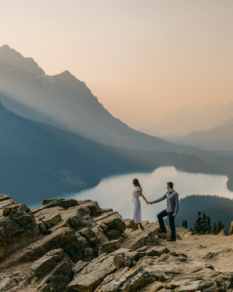 A breathtaking moment as a couple stands atop a mountain in Banff National Park by Moraine Lake with the bride’s dress flowing in the wind on a warm summer day. A dramatic photograph in the Canadian Rockies best elopement photographer captures the essence of adventure, love, and epic romance in the Canadian Rockies.