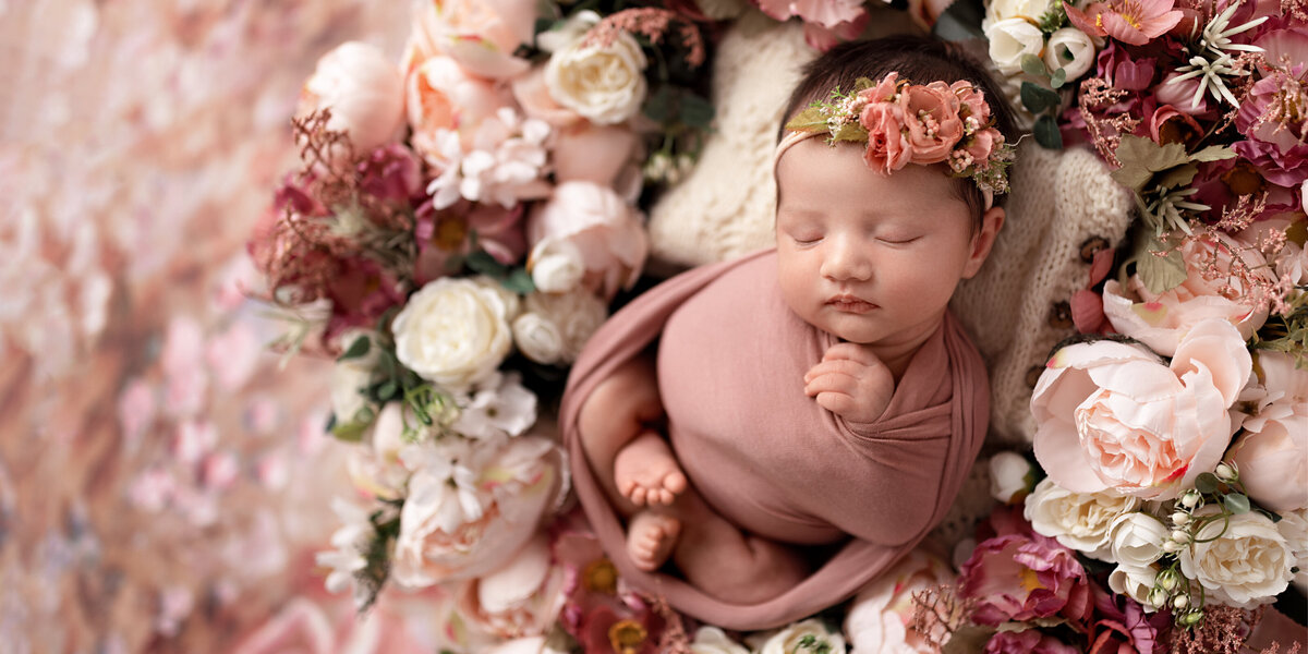 Newborn baby girl wrapped in pink and surrounded by soft pink and cream flowers, wearing a matching floral headband while sleeping peacefully.