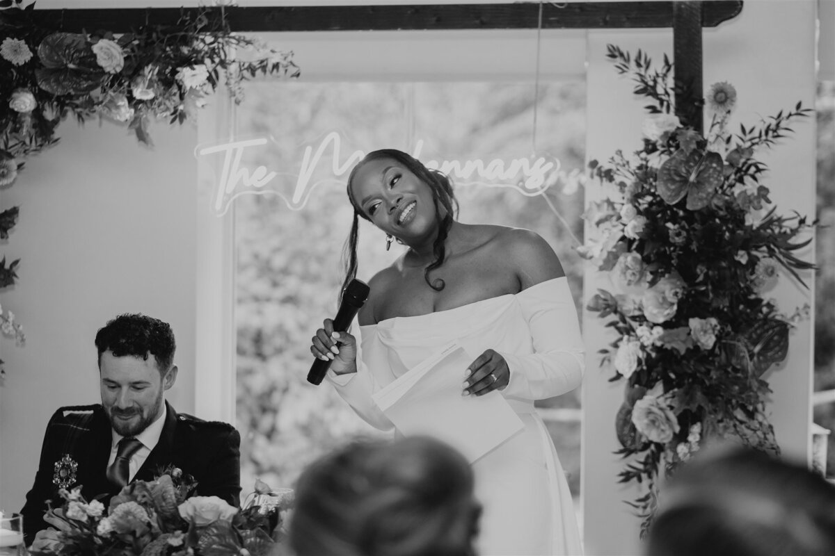 A bride stands in front of a window and faces a room full of seated wedding guests. She hold a microphone and delivers a wedding speech.