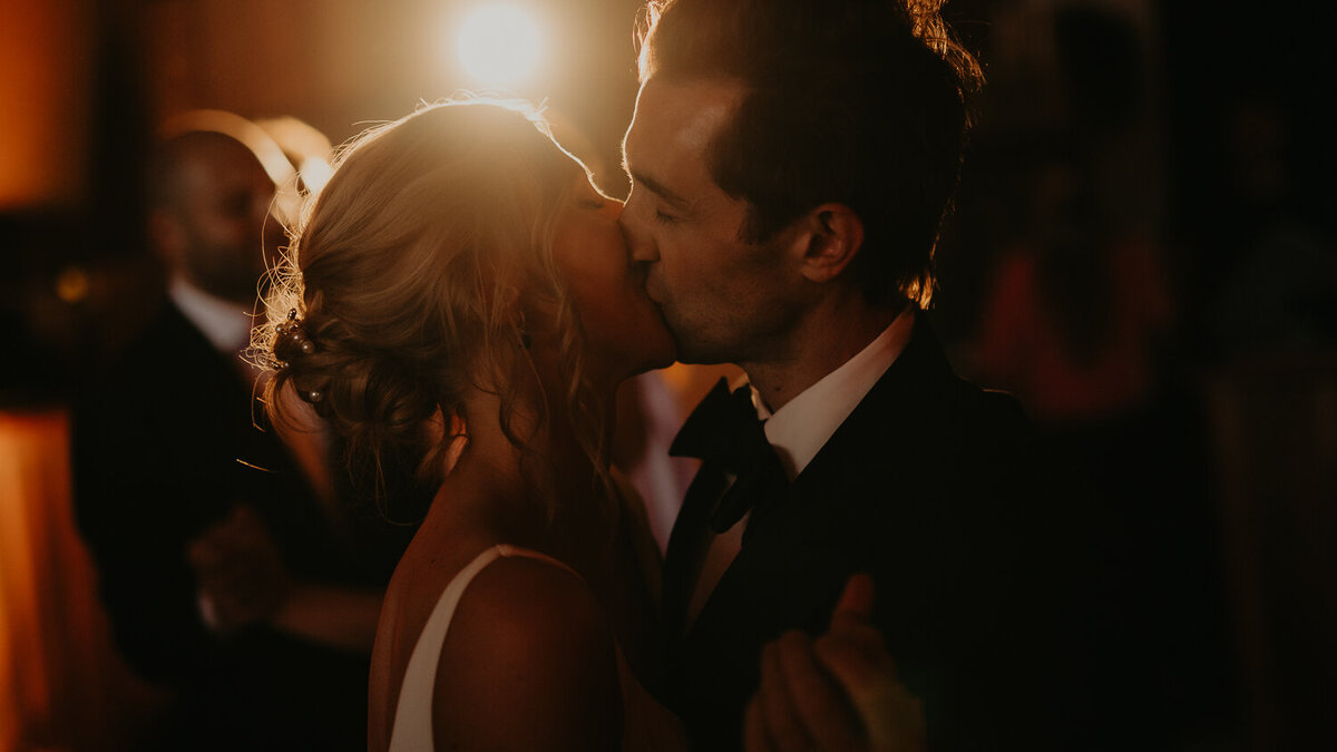 A bride and groom kiss during their first dance whilst being lit by an orange light at AMP Studios in London. 
