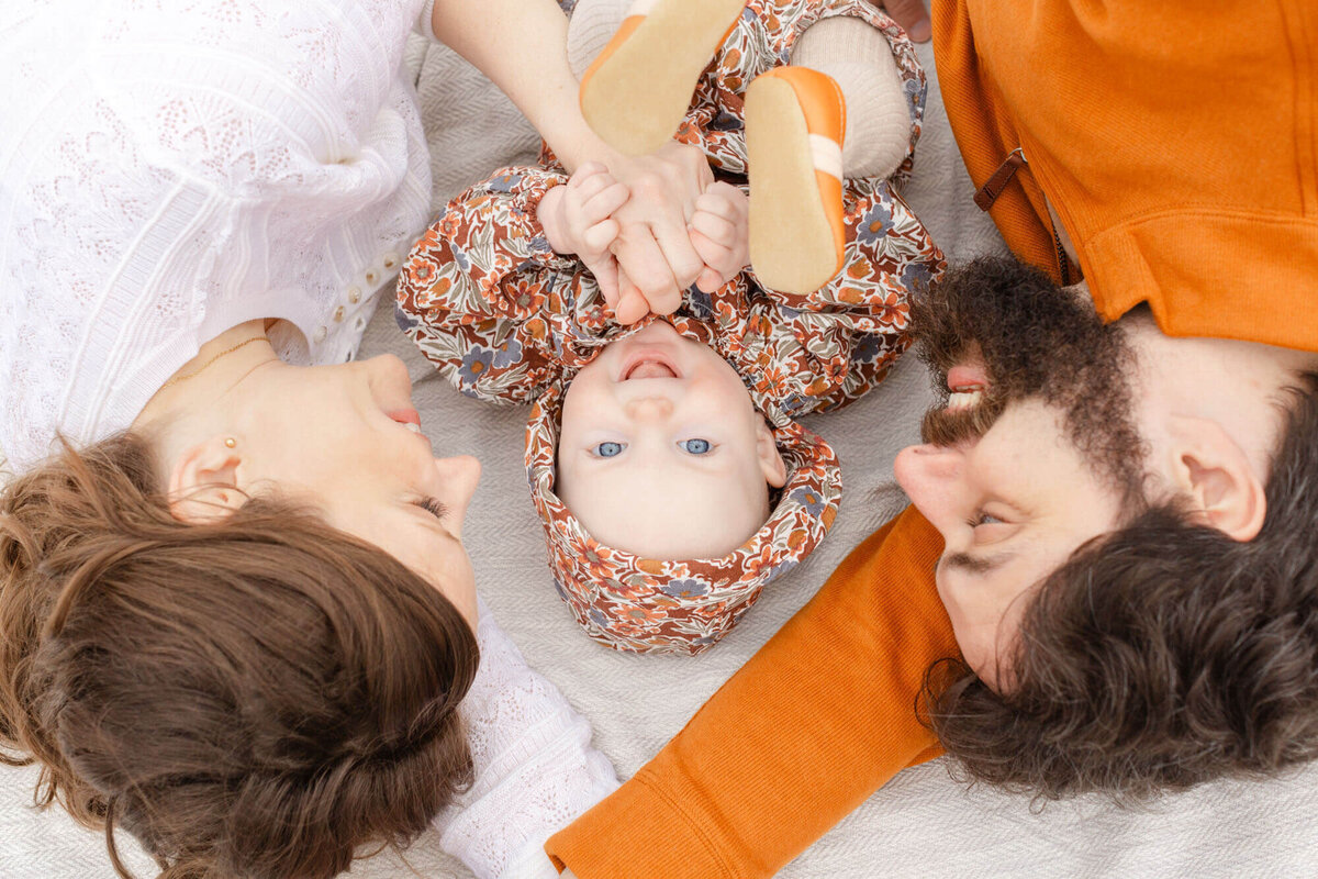 Baby is laying down on a blanket looking up at the camera. She is smiling and wearing a fall hued floral romper. Mom and dad are lying on either side of her and both are looking at her with big smiles on their faces.