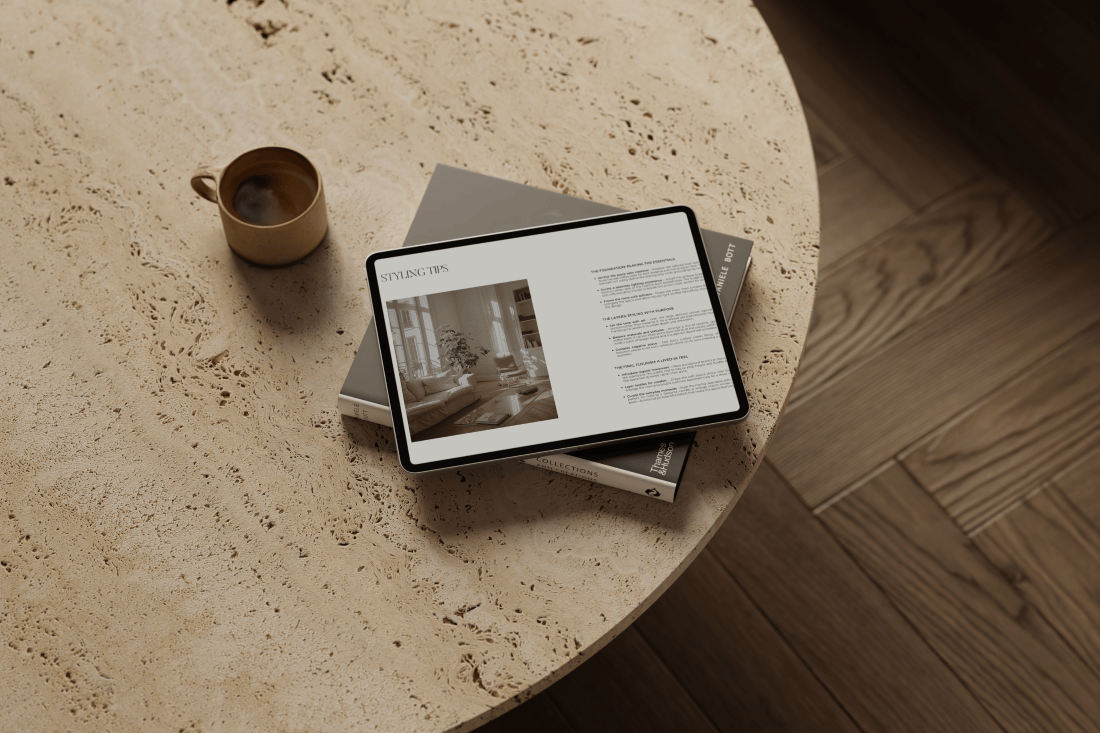A round travertine coffee table sits atop herringbone timber flooring, adorned with a coffee cup, an interior design book, and an iPad displaying the Styling Tips section from a bespoke Final Styling Scheme.