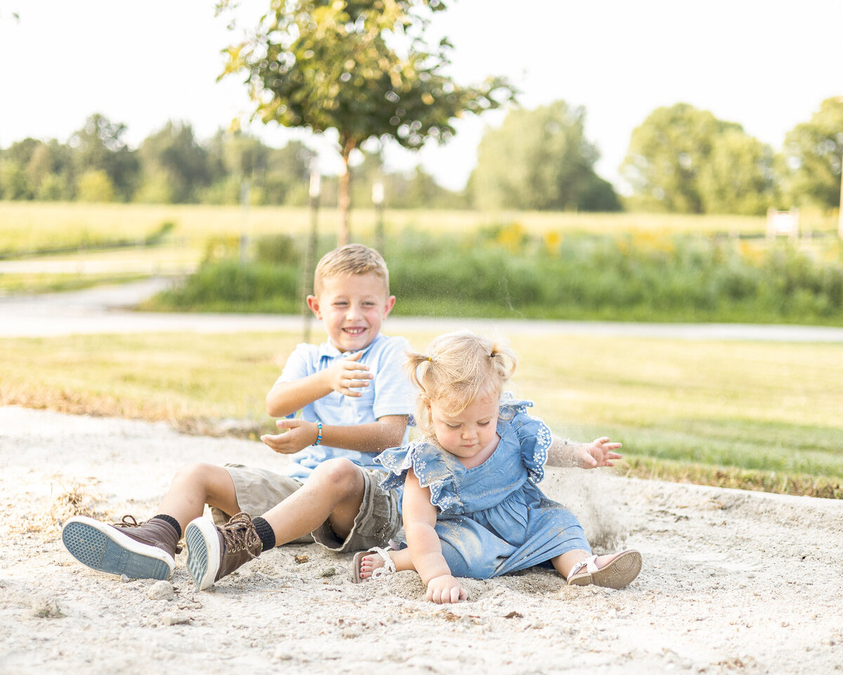 Young brother and sister sitting on the ground playing.  brother has short brown hair, wearing a partial button up short sleeve shirt, khaki shorts, brown shoes, smiling and throwing dirt at his sister.  Sister has blonde hair in pigtails, wearing a blue dress, white sandals, and looking down at the ground
