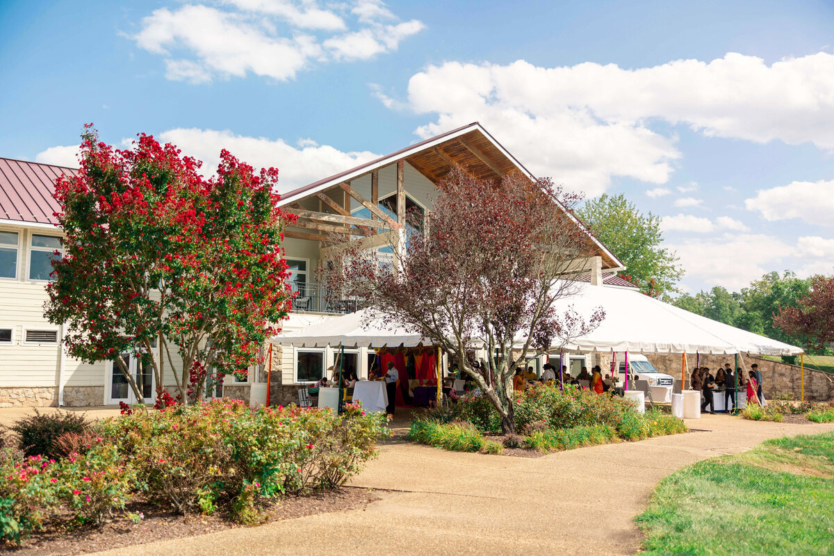A white tent set up beside a modern building with large windows on a sunny day. The scene includes red flowering trees, green bushes, and a clear blue sky with scattered clouds. People can be seen mingling under the tent.
