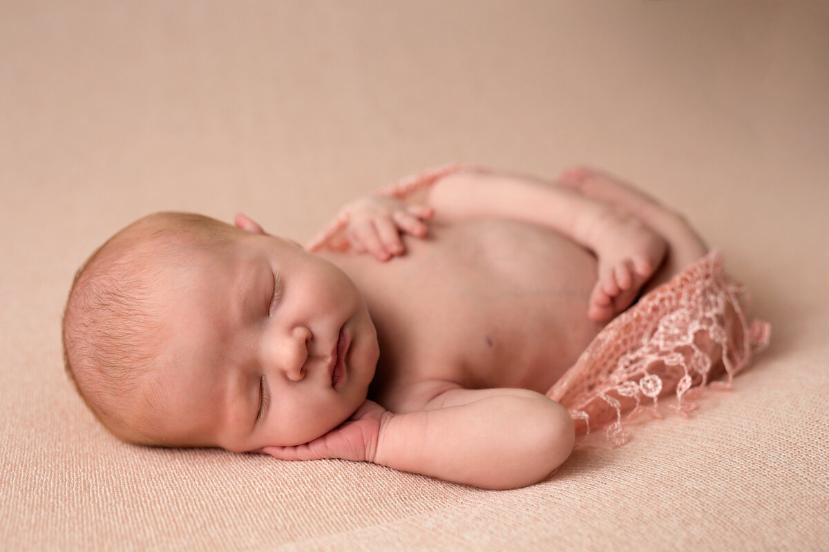 Baby girl with ginger hair laying on a peach background.  She is wrapped in peach lace near her legs.  She is asleep with her hand under her cheek and the other hand on her stomach.  Her legs are crossed.