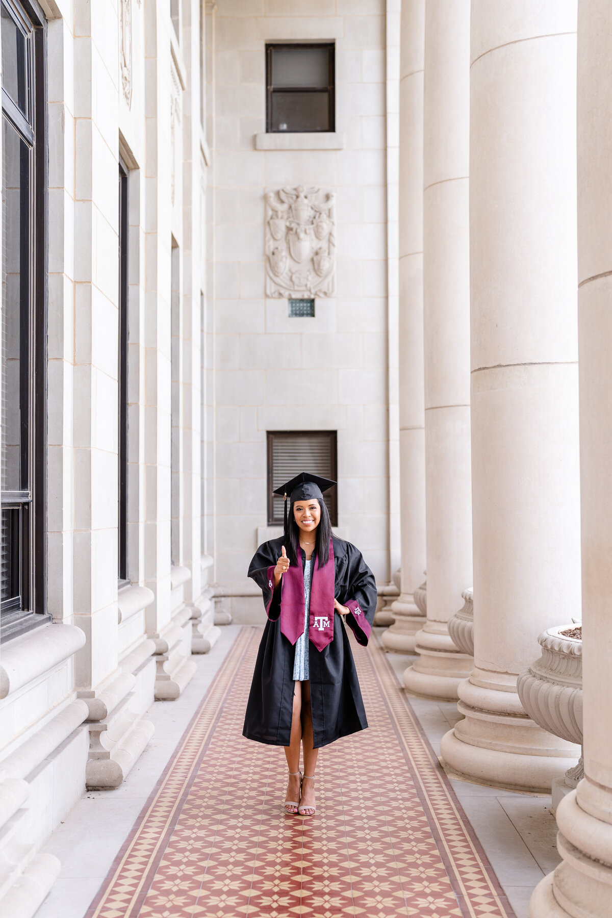 Texas A&M senior girl wearing cap, gown and stole and smiling with hand on hip and thumbs up in the columns of the Administration Building