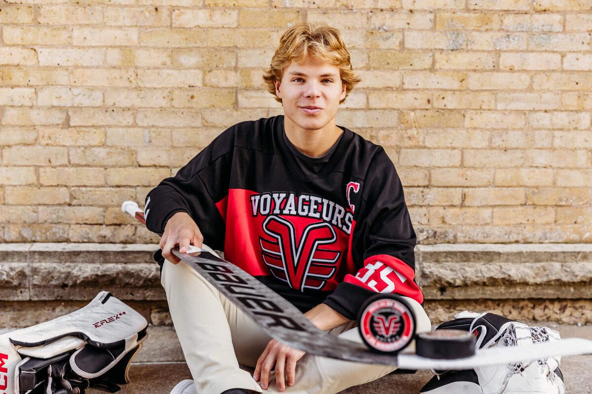 young man in his hockey jersey sitting against a brick wall and holding a hockey stick and balancing a puck on it for senior photos in Downtown Oshkosh