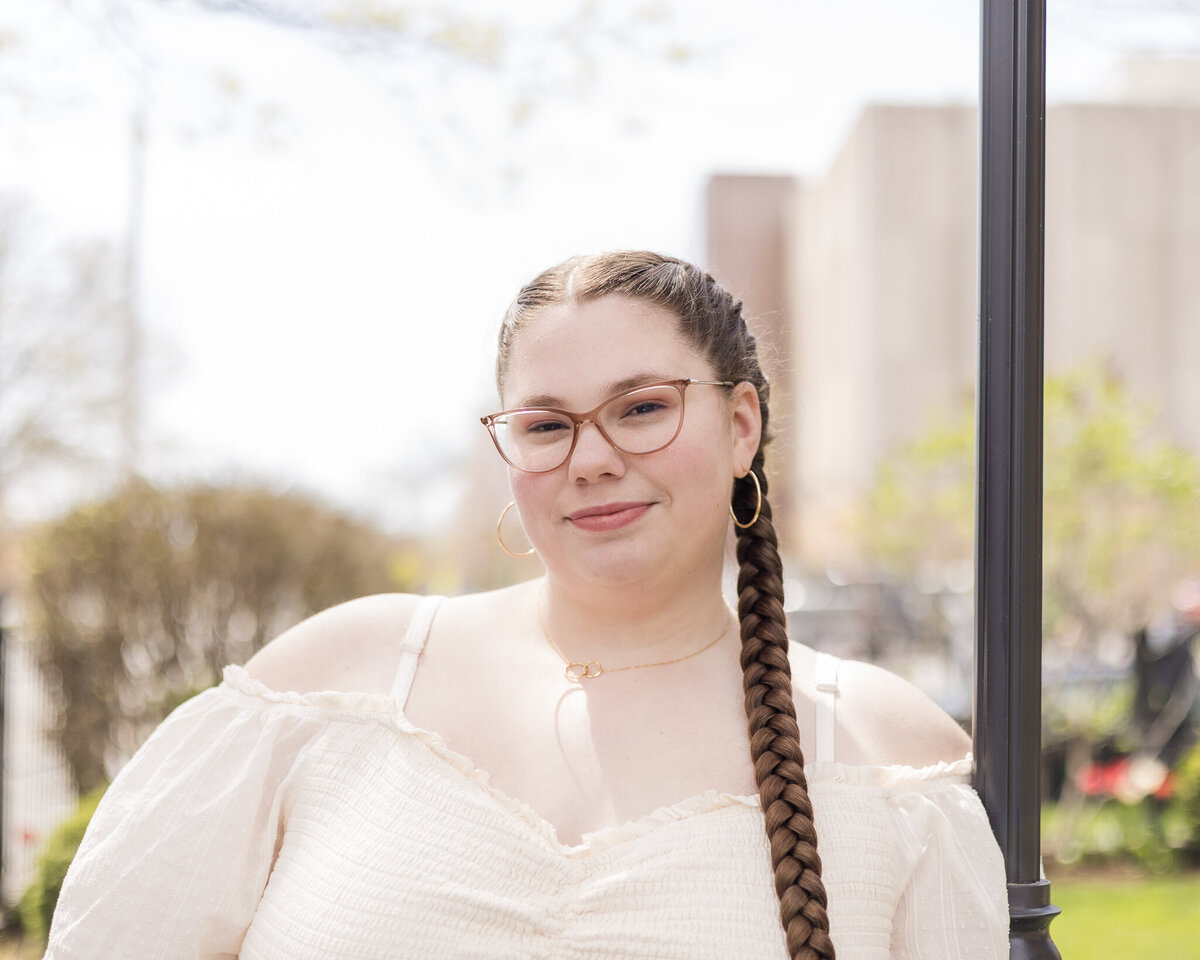 High school senior female with long dark brown braided hair, wearing a light creme shirt, glasses, standing next to a lamp post, smiling softly at the camera
