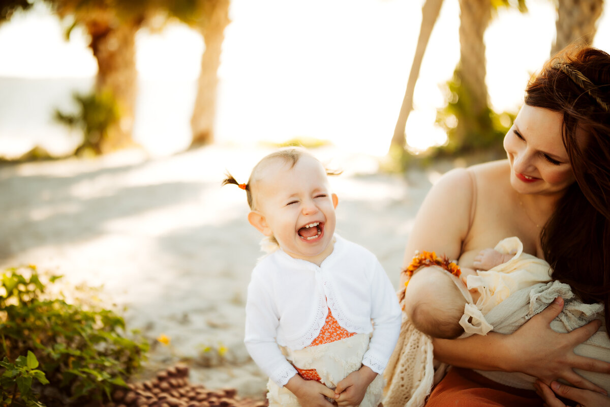 a toddler girl laughing hysterically white her mother breastfeeds her baby sister at the beach