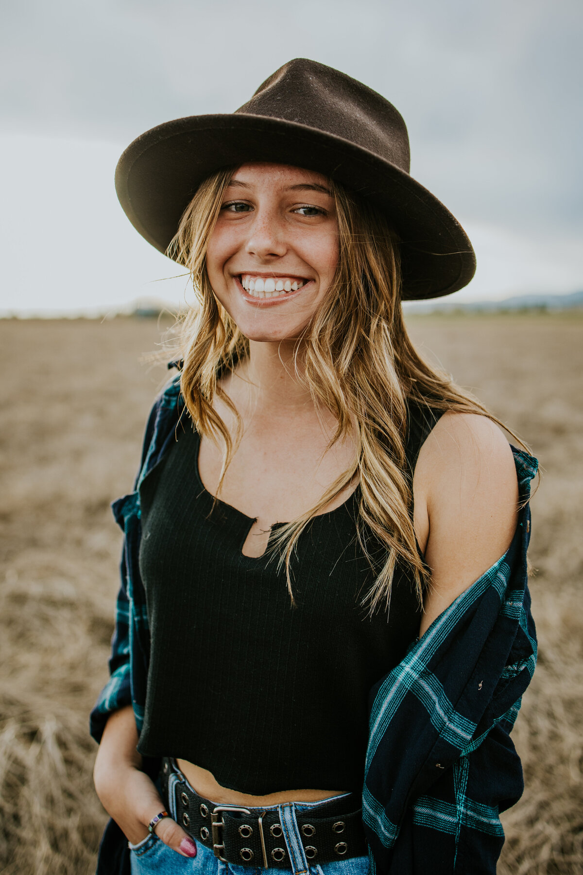 Young girl smiles at camera while wearing fedora in field.