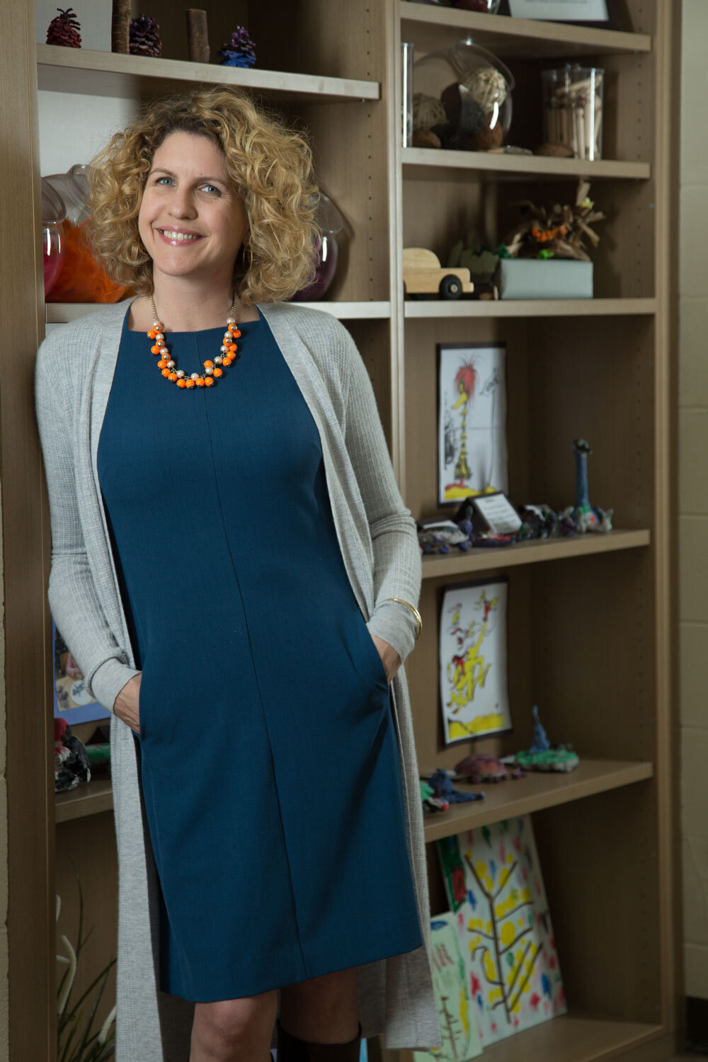 Woman in blue dress and gray sweater smiling in front of children's artwork