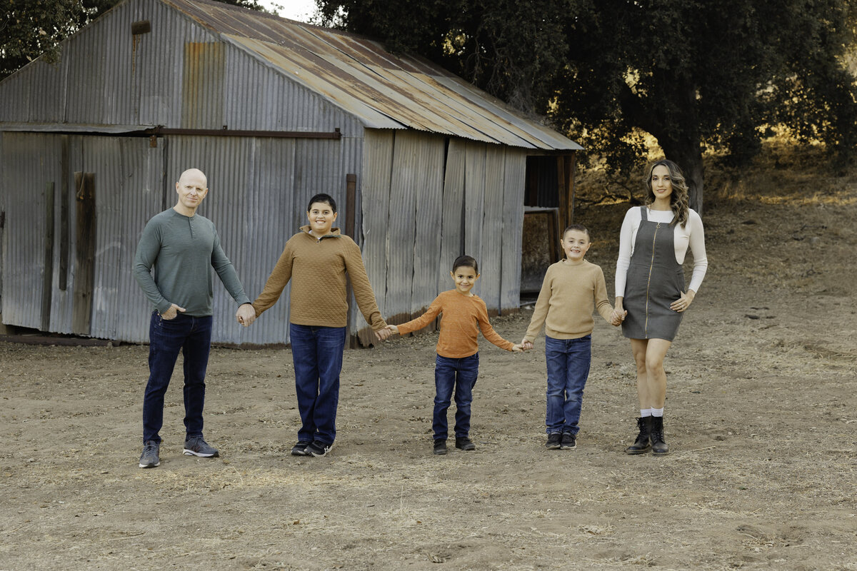 Family walking hand in hand in front of rustic barn backdrop, dressed in earthy tones. Photographed by Yucaipa family photographer Kaitlyn Dawn Photography.