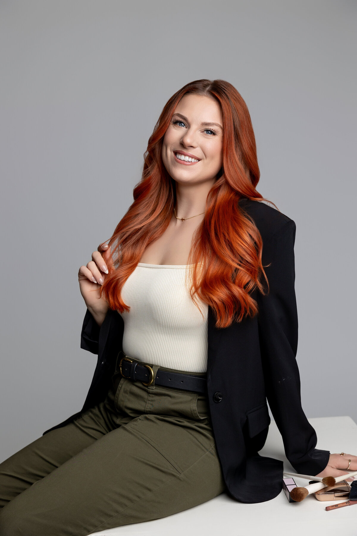 Woman with long red hair in black blazer holding compact mirror and makeup brush in studio. Photograph by Yucaipa branding photographer Kaitlyn Dawn Photography.