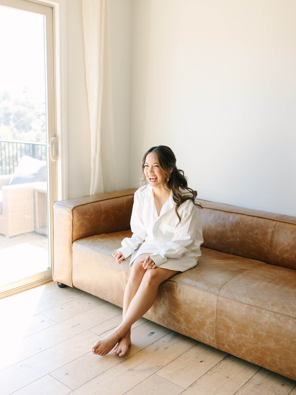 A bride in a white shirt sits joyfully on a tan leather couch, bathed in soft sunlight from a large window, exuding warmth.
